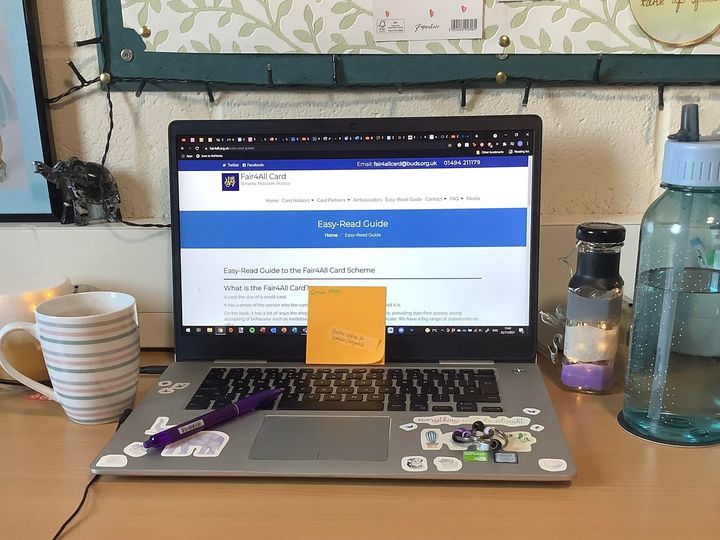 Image of a laptop with colourful stickers on, displaying a webpage reading easy-read guide. Next to the laptop are a water bottle and a mug. The laptop site on a brown desk, below a floral notice board.